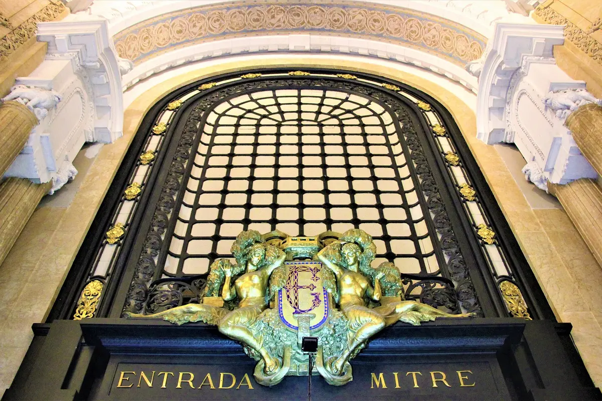 A tall arched iron window above a dark doorway marked Entrada Mitre, with two gilded bronze figures reclining beside a coat of arms, inside the Galería Güemes