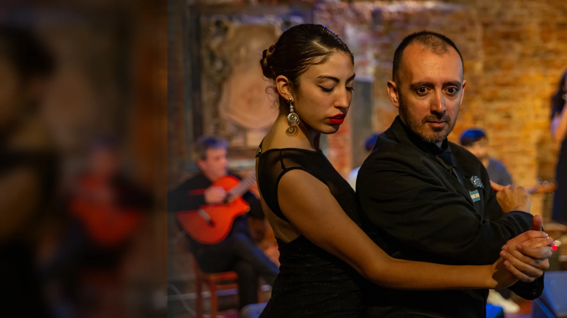 A tango couple in black dancing in close embrace while a guitarist and a singer perform behind them under warm light